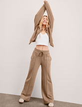 model wearing a brown zip-up sweatshirt, white cropped tank, and brown wide leg fleece pants against a plain background.
